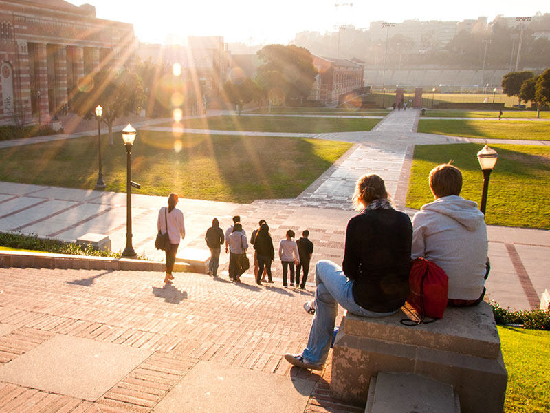 A couple dating in college sitting on a rock by the library watching the sun set on campus.