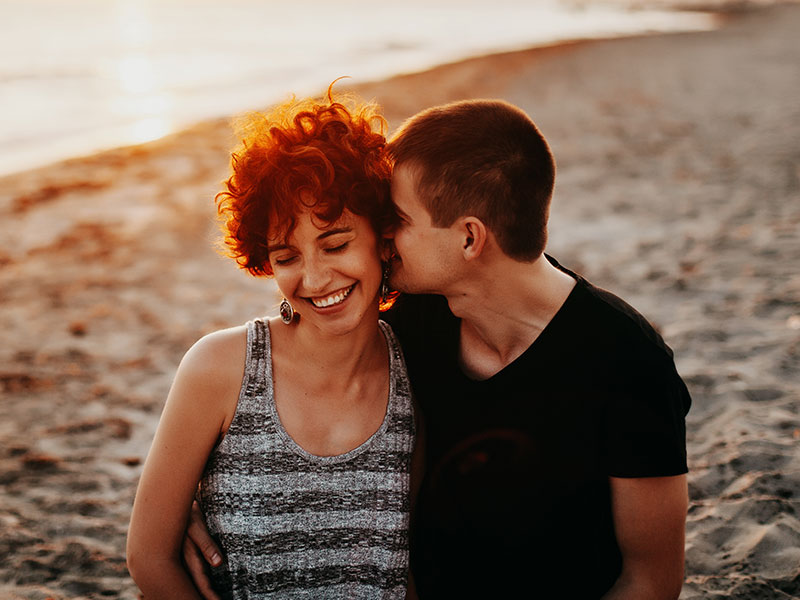 A woman who learned how to let your guard down, getting her cheek kissed on the beach while she laughs with her boyfriend.