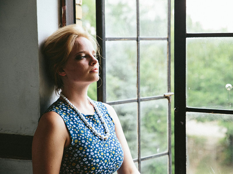 A woman who’s showing signs of low self-esteem leaning against the wall as she looks out the window looking resigned.