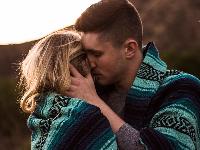 A couple of hopeless romantic people kissing at sunset while wrapped in a blanket.