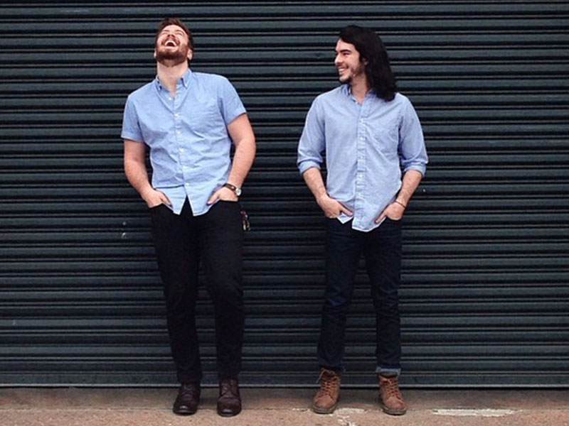 Two men who are life partners standing against a wall and laughing together in their engagement photos.