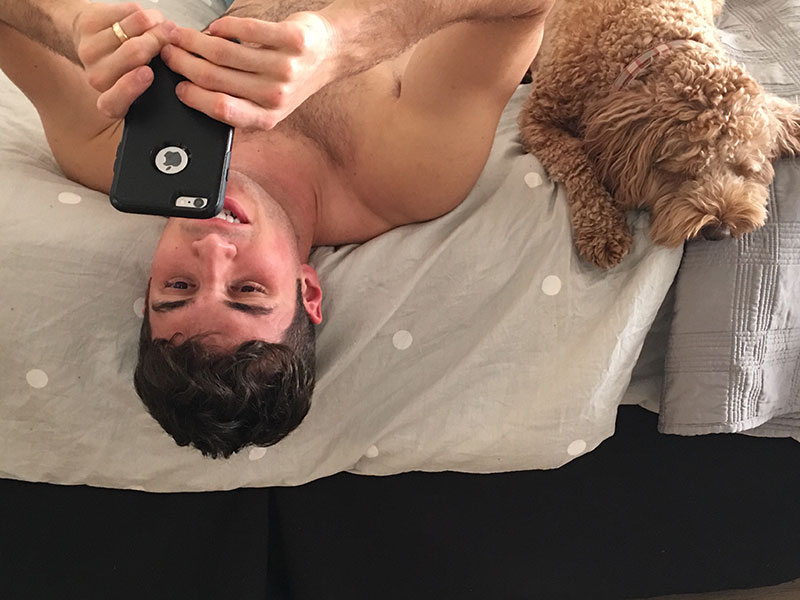 A guy writing Tinder messages while in bed with his dog next to him.
