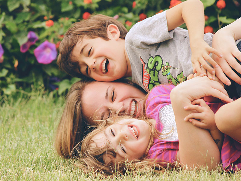 A woman who's dating a single parent with full custody laughing as she wrestles in the grass with two kids.