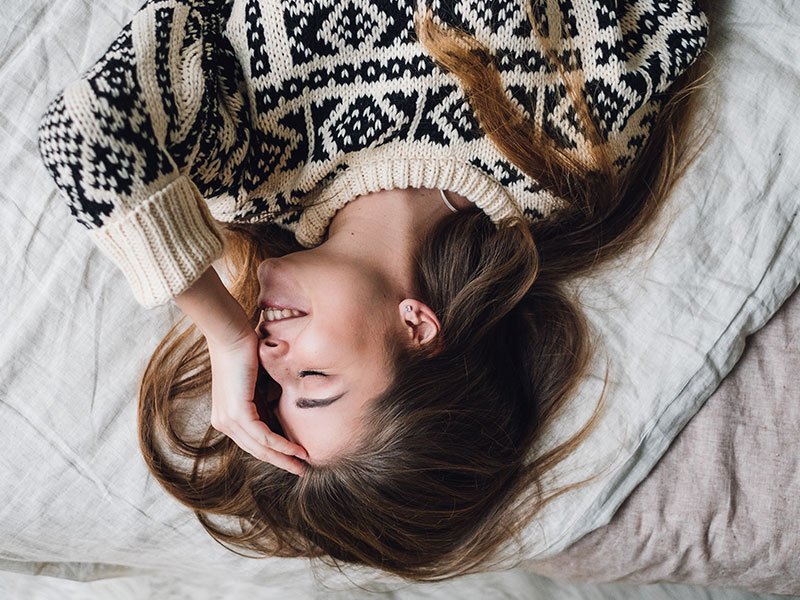 A woman going through a modern day breakup, on her bed laughing to herself with her hand on her head.