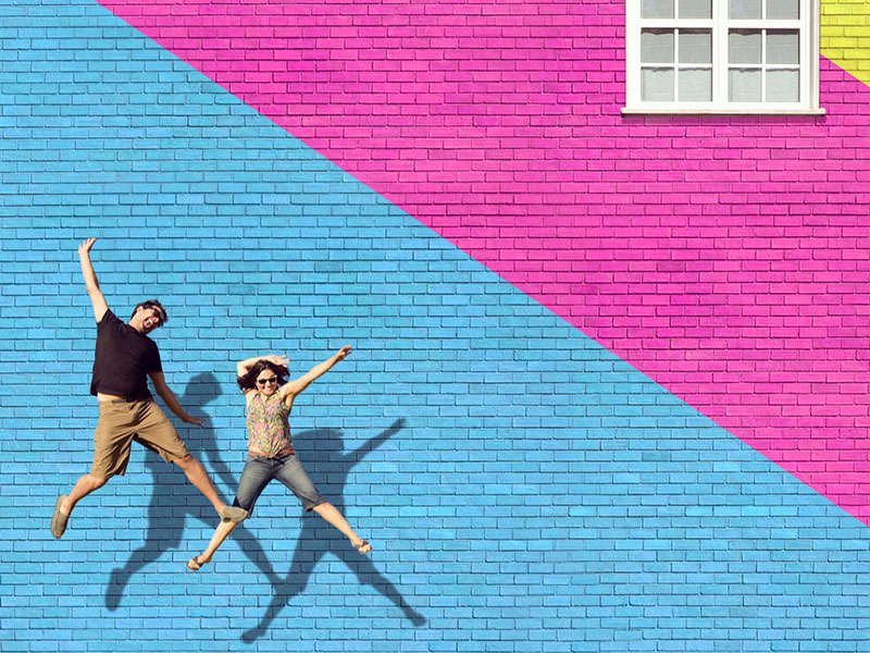 Two happy people who never settled in love, jumping in front of a colorful wall.