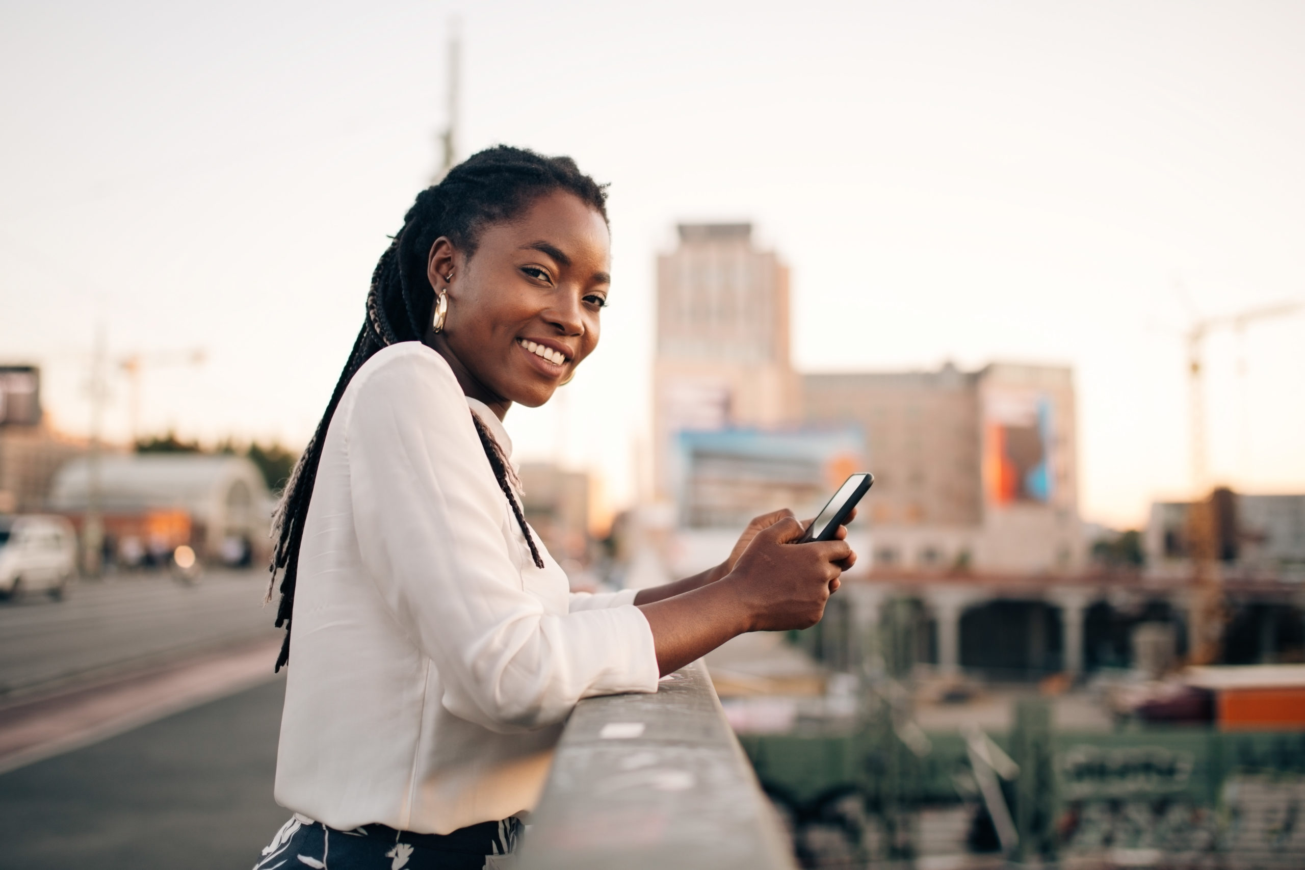 Young, smiling woman standing outdoors on a bridge and using mobile phone for Instagram dating.
