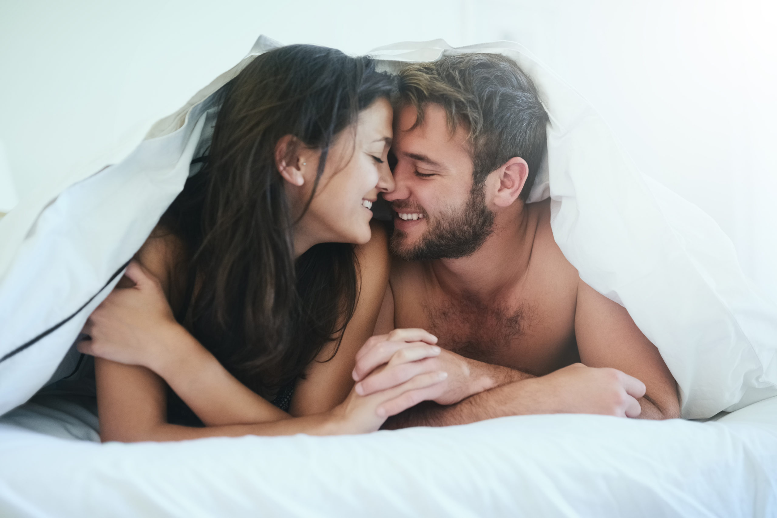Happy man and woman lying in bed together underneath the duvet and holding hands after enjoying the health benefits of sex.