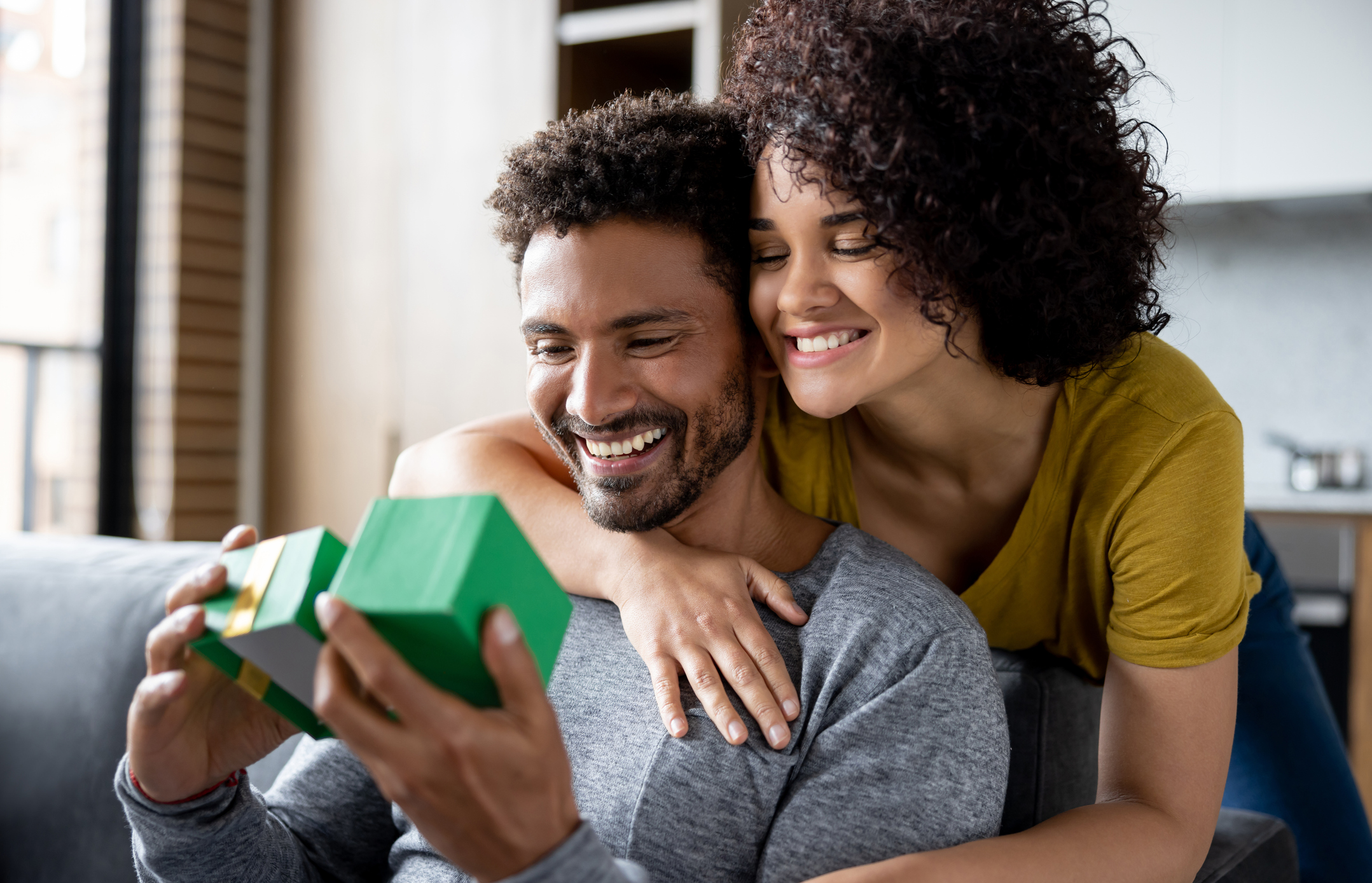 Smiling man with the gifts love language, sitting on the sofa opening a gift while girlfriend hugs him.