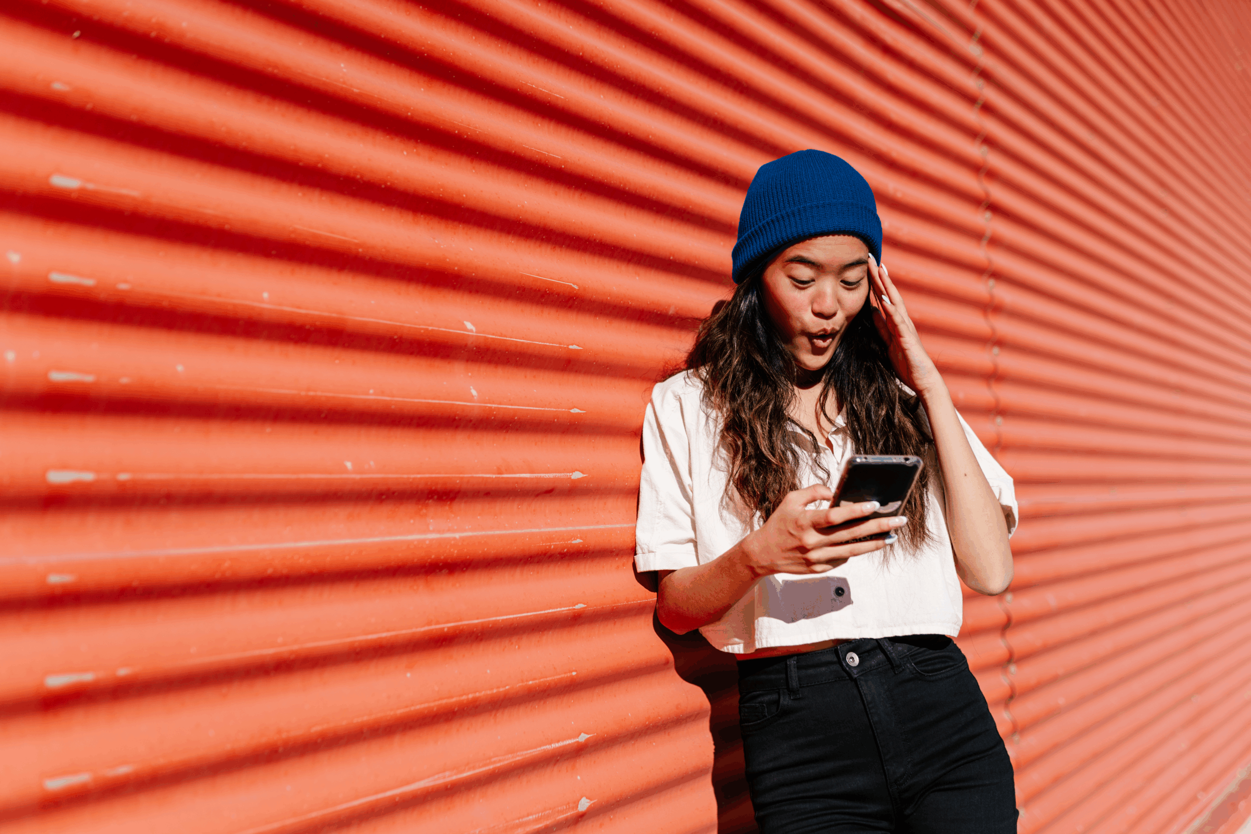 Young woman standing in front of red wall outdoors and smiling while using mobile phone and thinking about how to respond to pick-up lines.